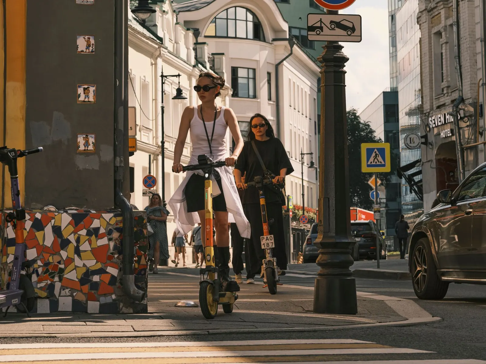 Two women riding electric scooters on a city street, showcasing urban commute.