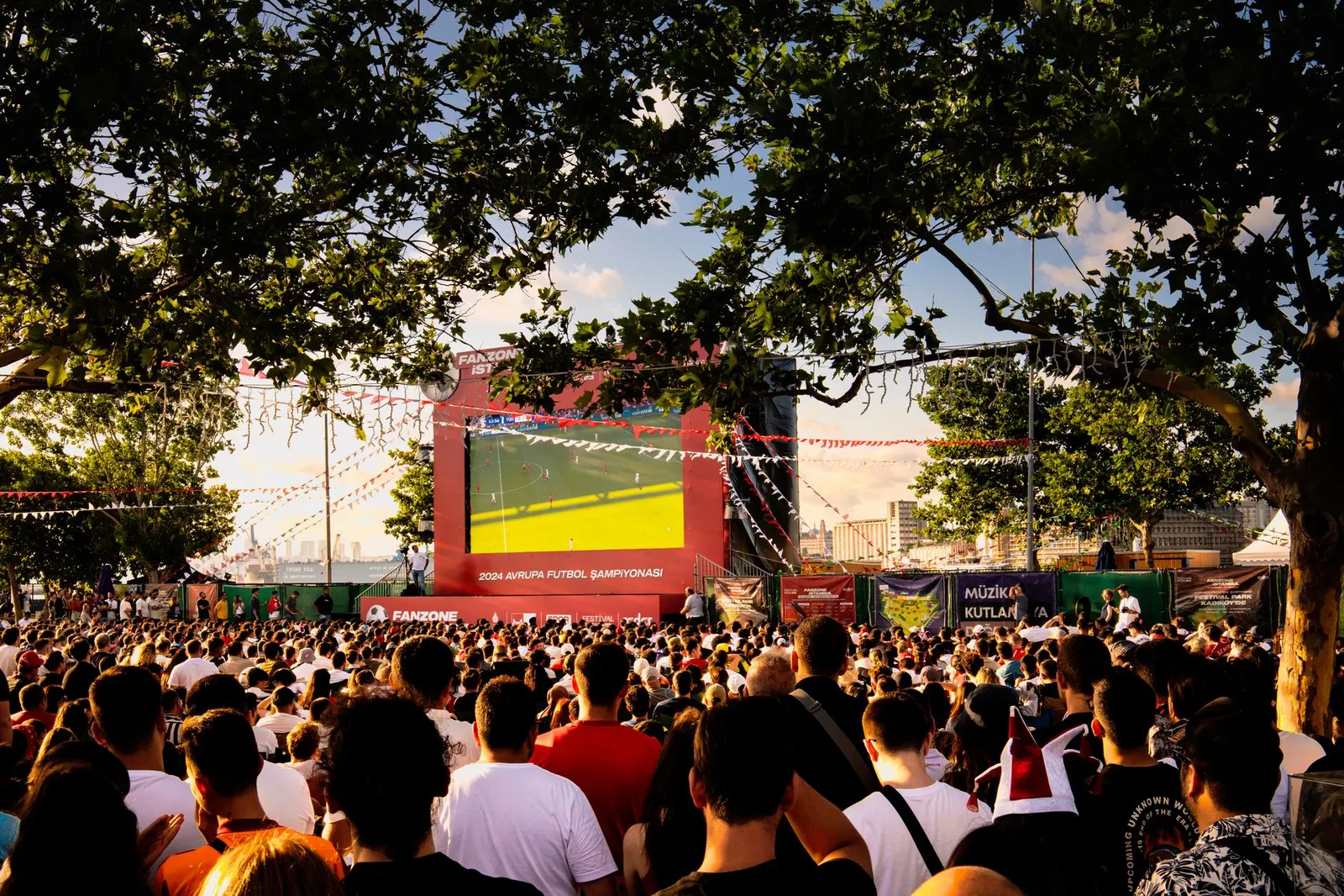 Crowd gathered in İstanbul park watching Euro football on big screen. Energetic summer atmosphere.